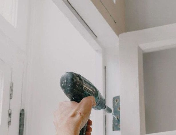 Back view of unrecognizable craftsman using cordless screwdriver and assembling window while screwing metal hinge during renovation works in apartment